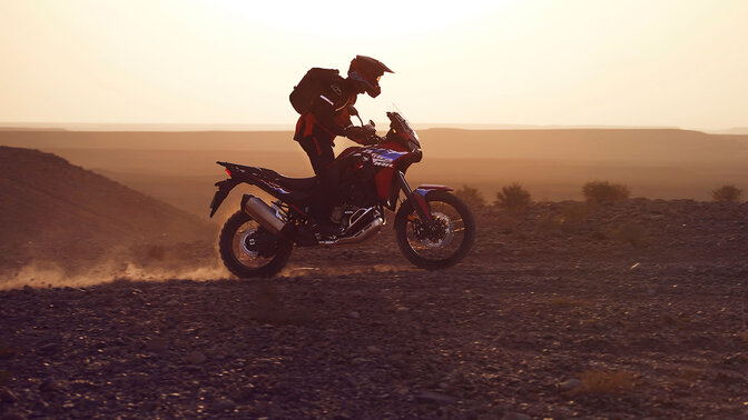 Model rijdend op een CRF1100L Africa Twin motorfiets op een woestijnlocatie.