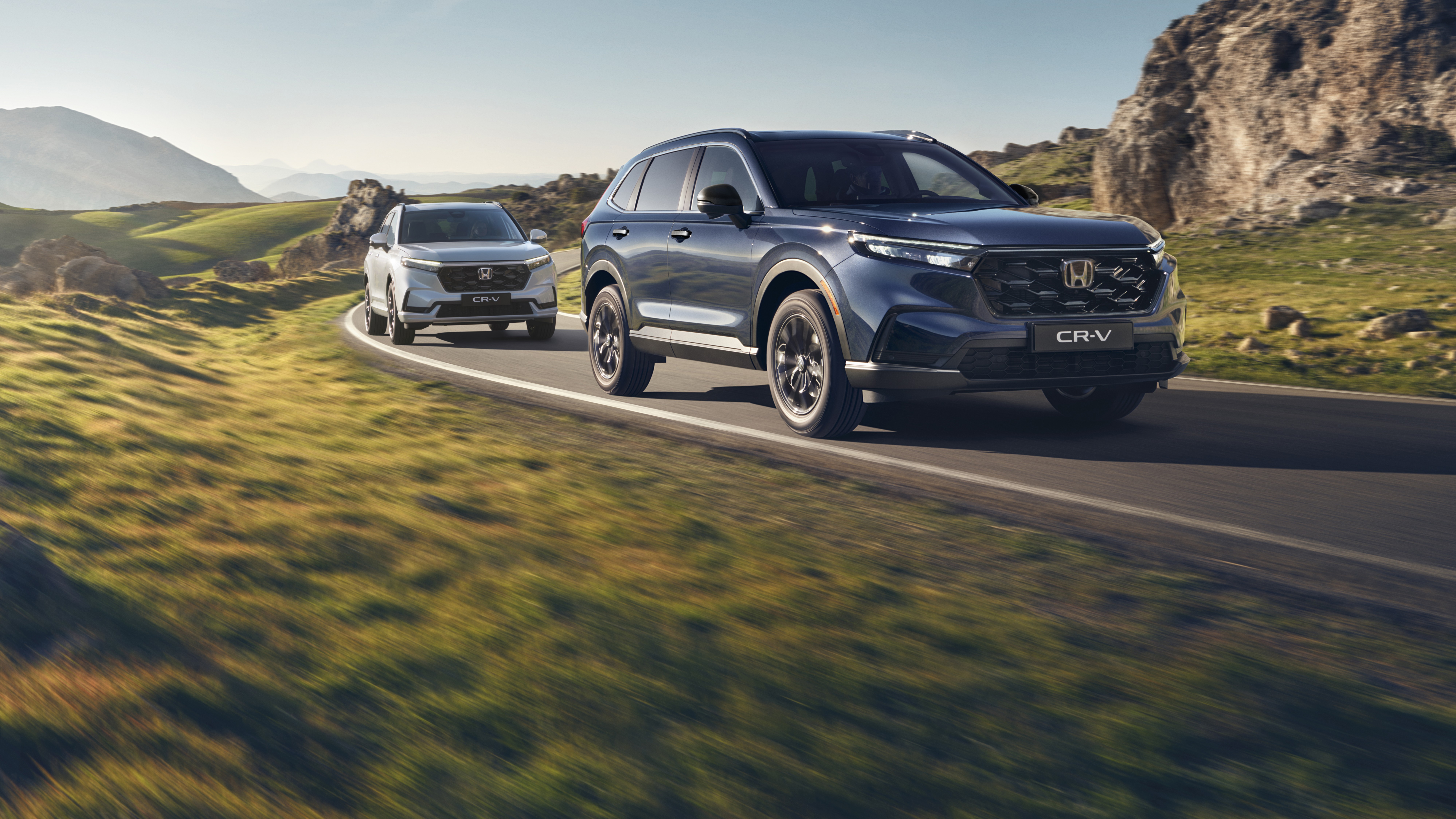A silver Honda CR-V SUV driving on a winding mountain road with rocky terrain and mountains in the background. The vehicle is in motion, creating a sense of speed and adventure in the rugged landscape.