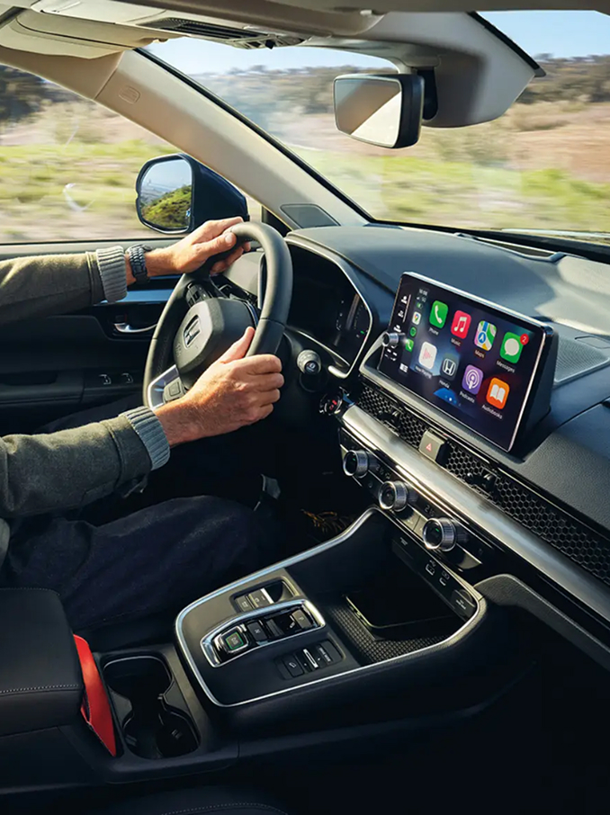 Interieur van een moderne auto met een bestuurder achter het stuur. Het dashboard, stuurwiel, middenconsole en een breed touchscreen display met kleurrijke app-iconen zijn zichtbaar, terwijl buiten de ramen het landschap voorbij trekt.