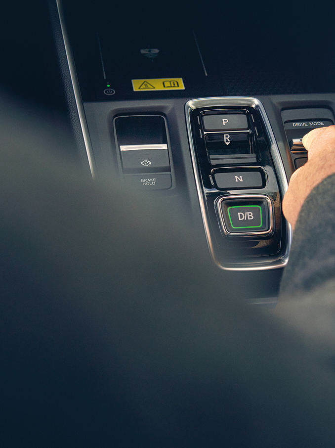Iemand reikt om een tuimelschakelaar op de middenconsole van een moderne auto te verstellen, vlak naast de versnellingsknoppen.