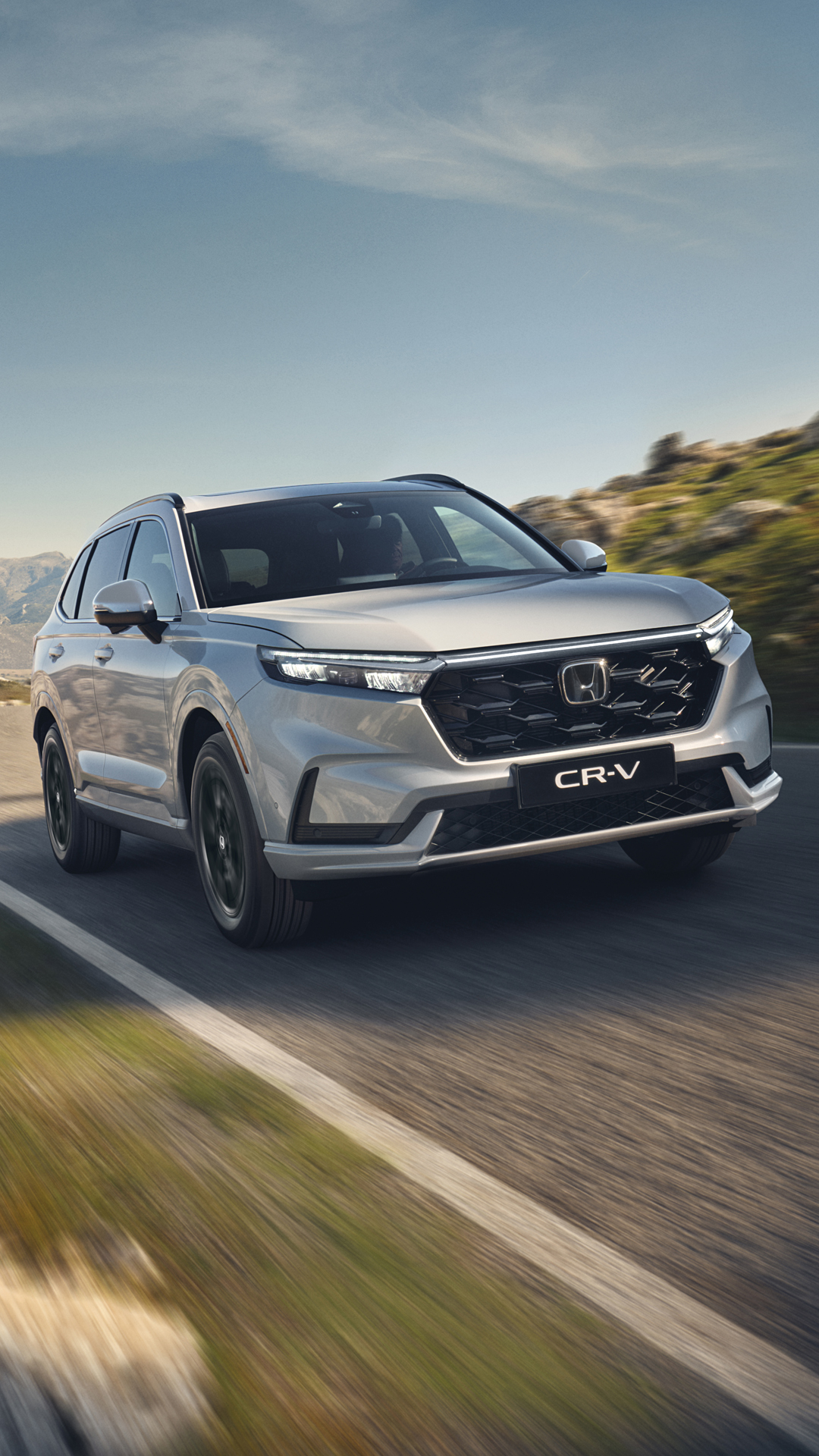 A silver Honda CR-V SUV driving on a winding mountain road. The vehicle is in motion, with blurred scenery visible in the background. The front grille and headlights of the new CR-V model are prominently displayed.