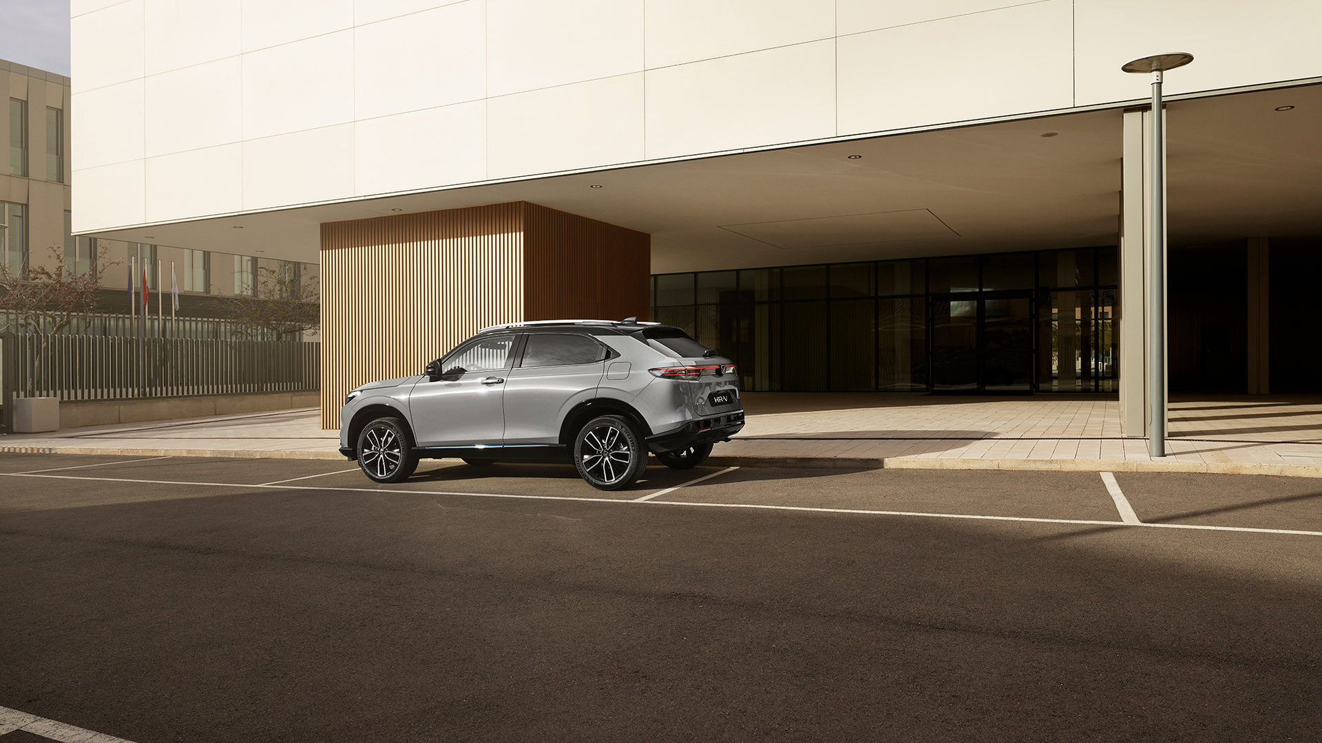 A silver Honda HR-V is parked in an outdoor parking space in front of a modern building with glass walls and vertical wooden paneling, photographed from a rear three-quarter angle in daylight.