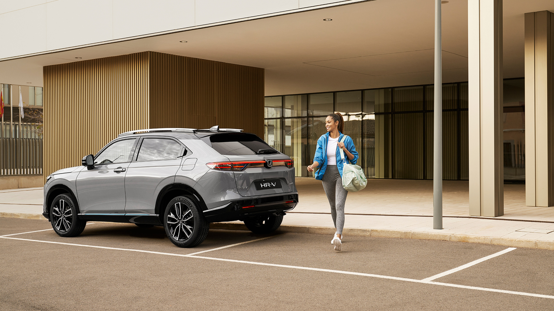 A silver Honda HR-V parked outside a modern building while a woman walks past it carrying a bag, suggesting everyday urban use and convenience.