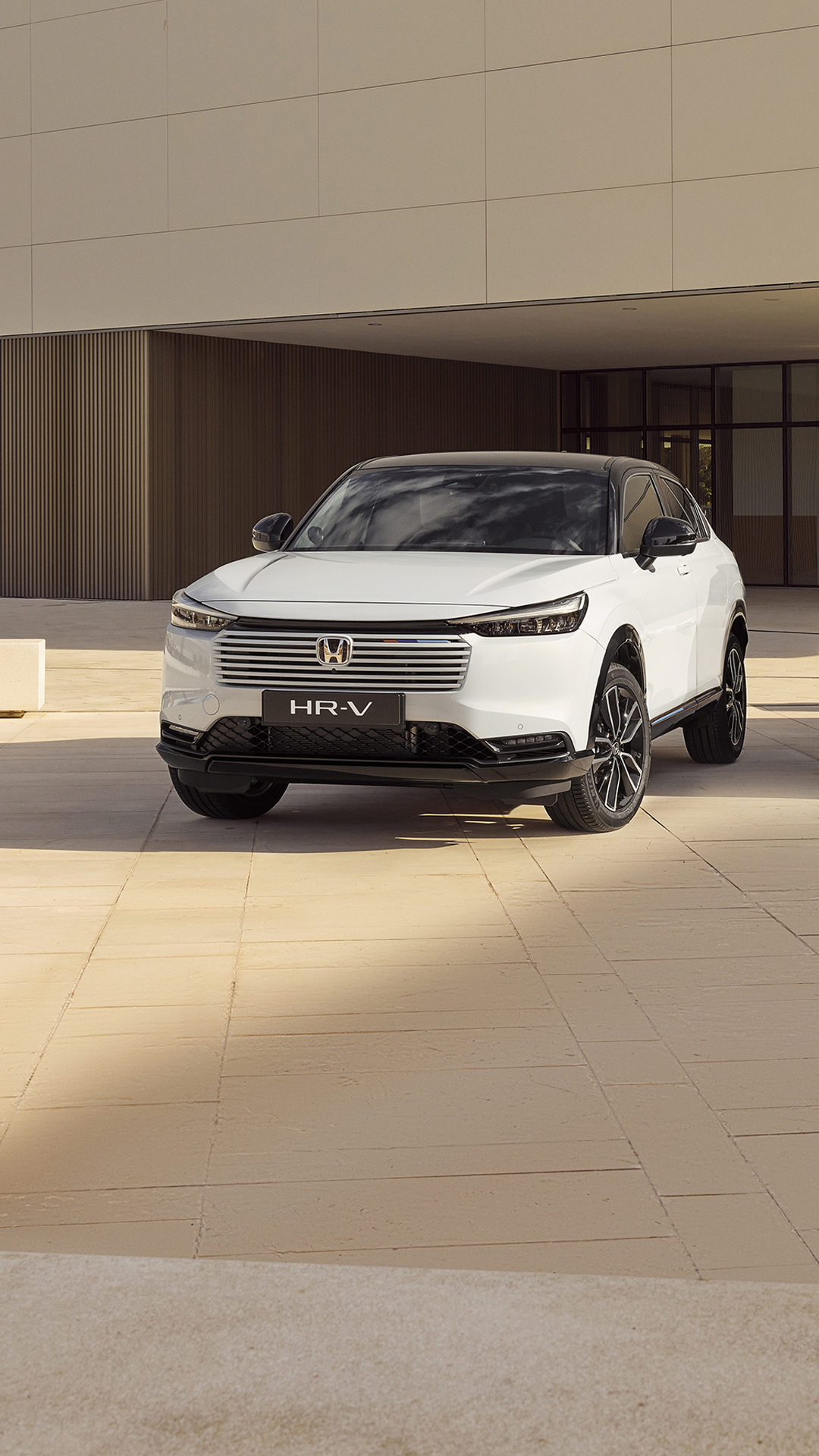 A white Honda HR-V SUV parked in front of a modern building with beige tiled walls. The vehicle is positioned at an angle, showcasing its sleek design and distinctive front grille.