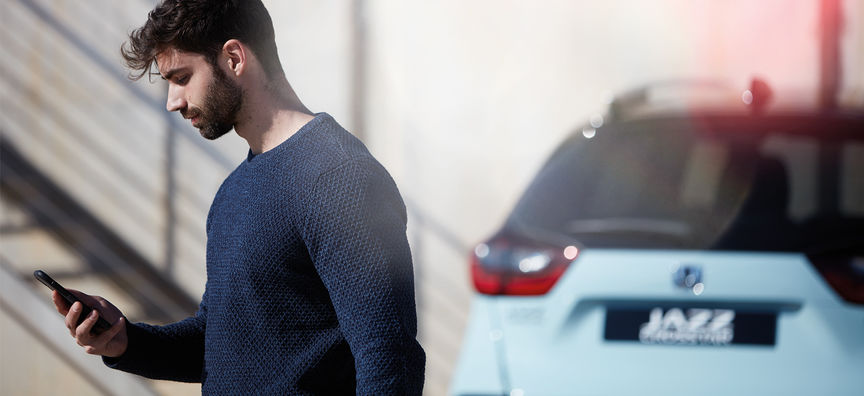 Een man kijkt naar de My Honda+ app met een Honda Jazz Hybrid Crosstar op de achtergrond. 