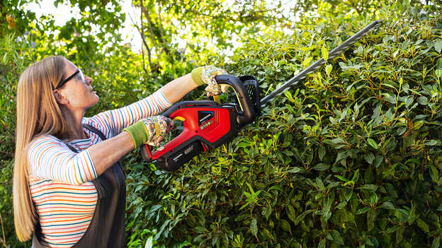 Vrouw trimt bosjes met accu Honda-heggenschaar in een tuin.