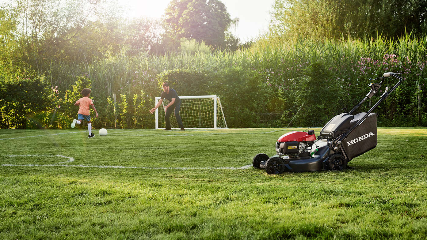 Man en kind voetballend naast zijwaarts gerichte Honda HRN in de tuin. 