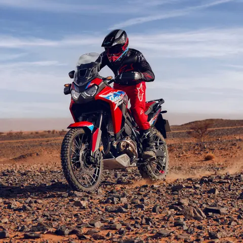 Model rijdend op een CRF1100L Africa Twin motorfiets op rotsachtig terrein op een woestijnlocatie.