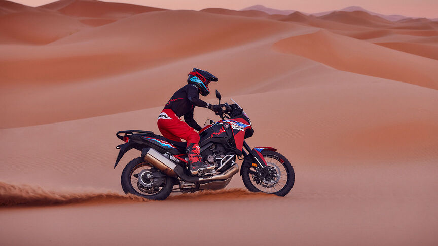 Model rijdend op een CRF1100L Africa Twin motorfiets op een woestijnlocatie.