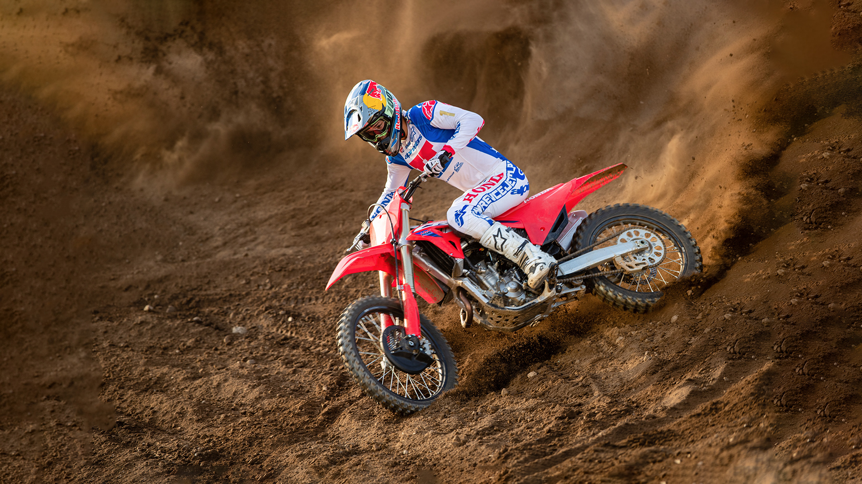 Een motorcrosser in witte, blauwe en rode race-uitrusting leunt diep in een bocht op een rode crossmotor, snijdt door een zanderig circuit en werpt een spectaculaire wolk zand achter zich op.