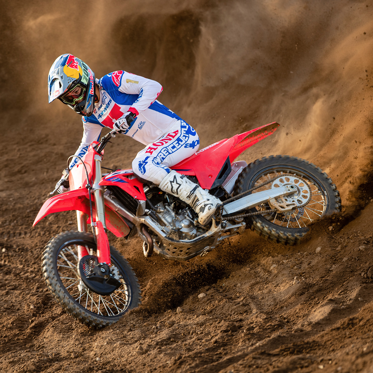 Een motorcrosser in witte, blauwe en rode race-uitrusting leunt diep in een bocht op een rode crossmotor, snijdt door een zanderig circuit en werpt een spectaculaire wolk zand achter zich op.