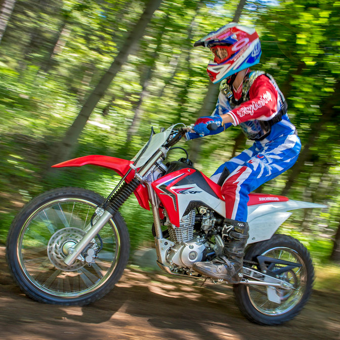 Zijaanzicht van CRF125F met rijder op onverharde weg op racelocatie in de bossen. Naar links.