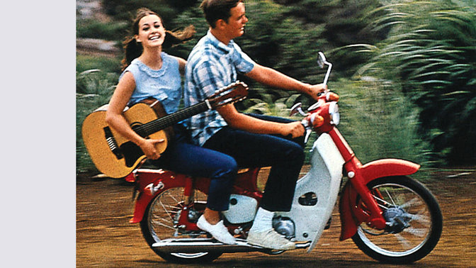 Honda Cub met rijder en passagier met een gitaar. Op een onverharde weg. Zicht op rechterkant.